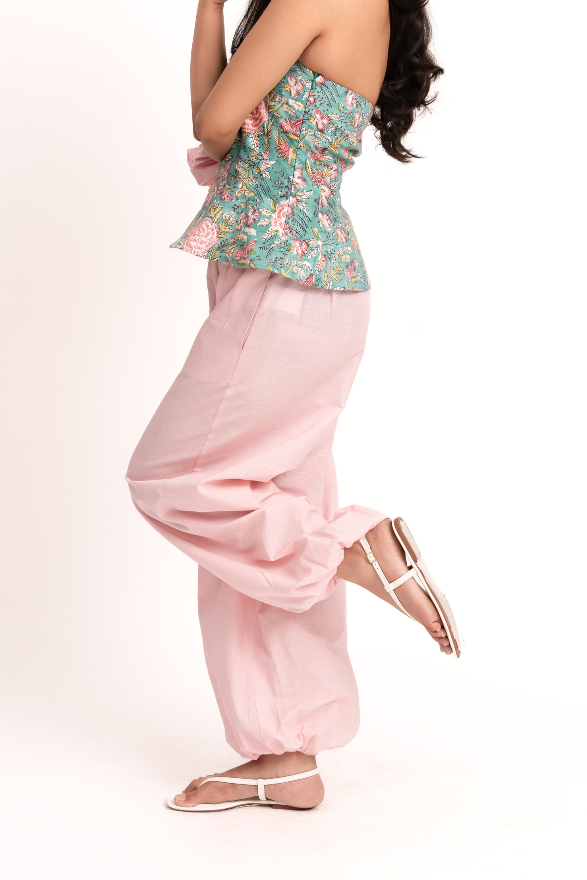 A woman modeling a strapless peplum top with a green tropical floral print and matching pink wide-leg pants, paired with white strappy flat sandals, against a plain white studio background.