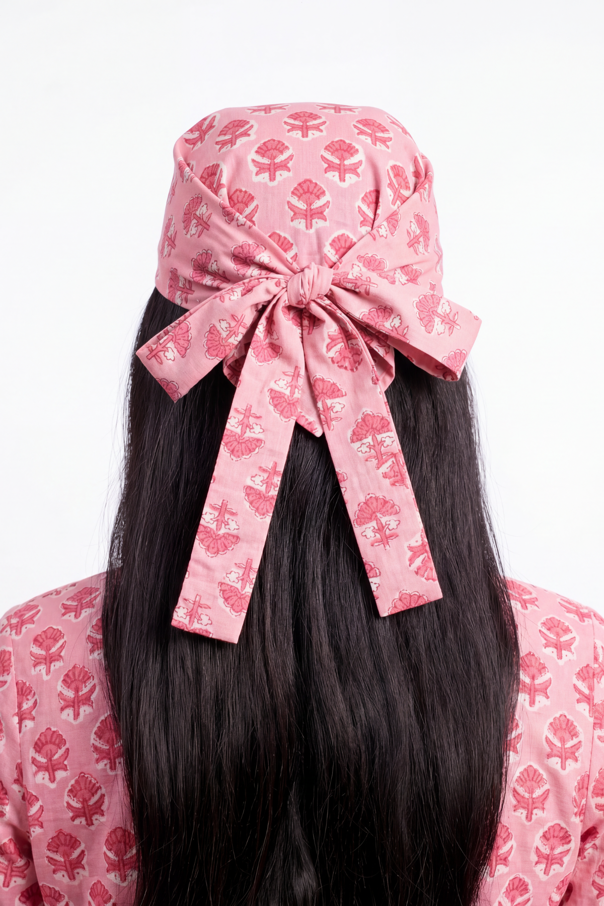 A pink fabric headscarf with a matching bow, featuring a red floral block print, styled on a model with long black hair against a white background.