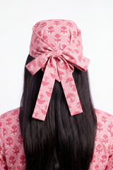 A pink fabric headscarf with a matching bow, featuring a red floral block print, styled on a model with long black hair against a white background.