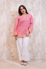 A woman modeling a pink long-sleeve kurta with a yellow and white geometric pattern and yellow border trim, paired with white wide-leg pants and white sandals, standing against a textured beige wall.