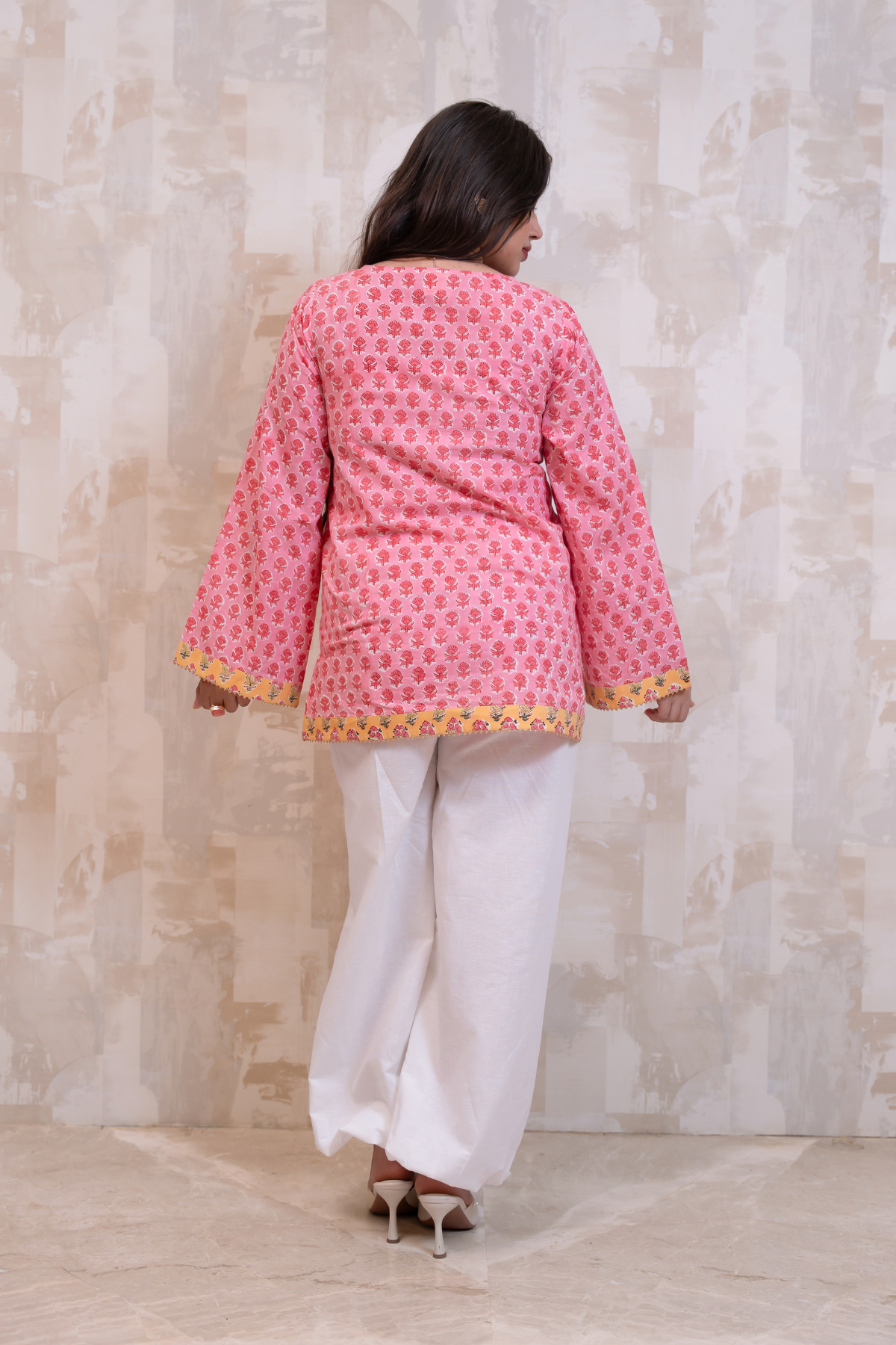 A woman viewed from behind wearing a pink printed kurta with yellow and orange patterned lace trim, paired with white wide-leg trousers and beige heels, standing against a textured beige wall.