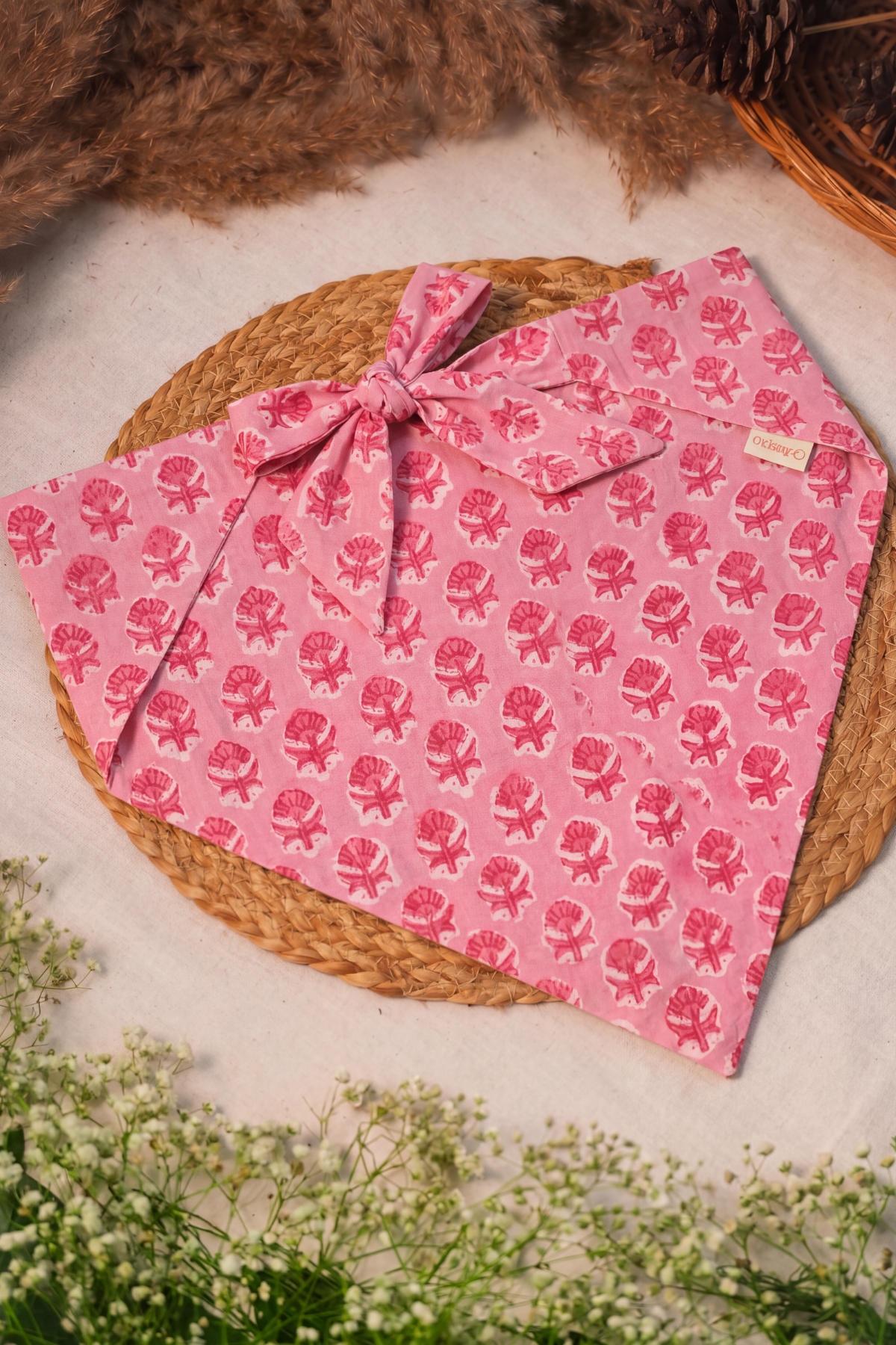 A pink fabric bandana with a repeating pink floral block print pattern and a tied bow, styled on a round woven straw mat with dried brown grass and small white flowers.