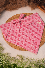 A pink fabric bandana with a repeating pink floral block print pattern and a tied bow, styled on a round woven straw mat with dried brown grass and small white flowers.
