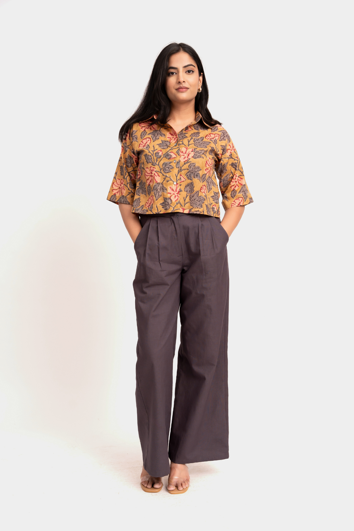 A woman modeling a short-sleeve button-up shirt with a warm-toned floral pattern in orange, yellow, and brown, paired with dark charcoal gray wide-leg trousers, against a plain light gray studio background.