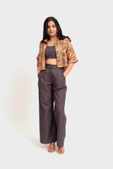 A woman modeling a two-piece outfit consisting of a dark brown crop top and matching wide-leg trousers, paired with a short-sleeved cropped floral jacket in orange, yellow, and brown tones, set against a plain light gray background.