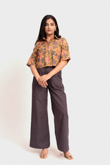 A woman modeling a short-sleeve button-up shirt with a colorful orange, yellow, and green floral pattern, paired with dark gray wide-leg trousers, against a light gray studio background.