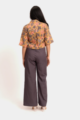 A woman shown from the back wearing a short-sleeve collared button-up shirt with a yellow, orange, and gray floral pattern, paired with wide-leg dark gray trousers, against a light gray studio background.