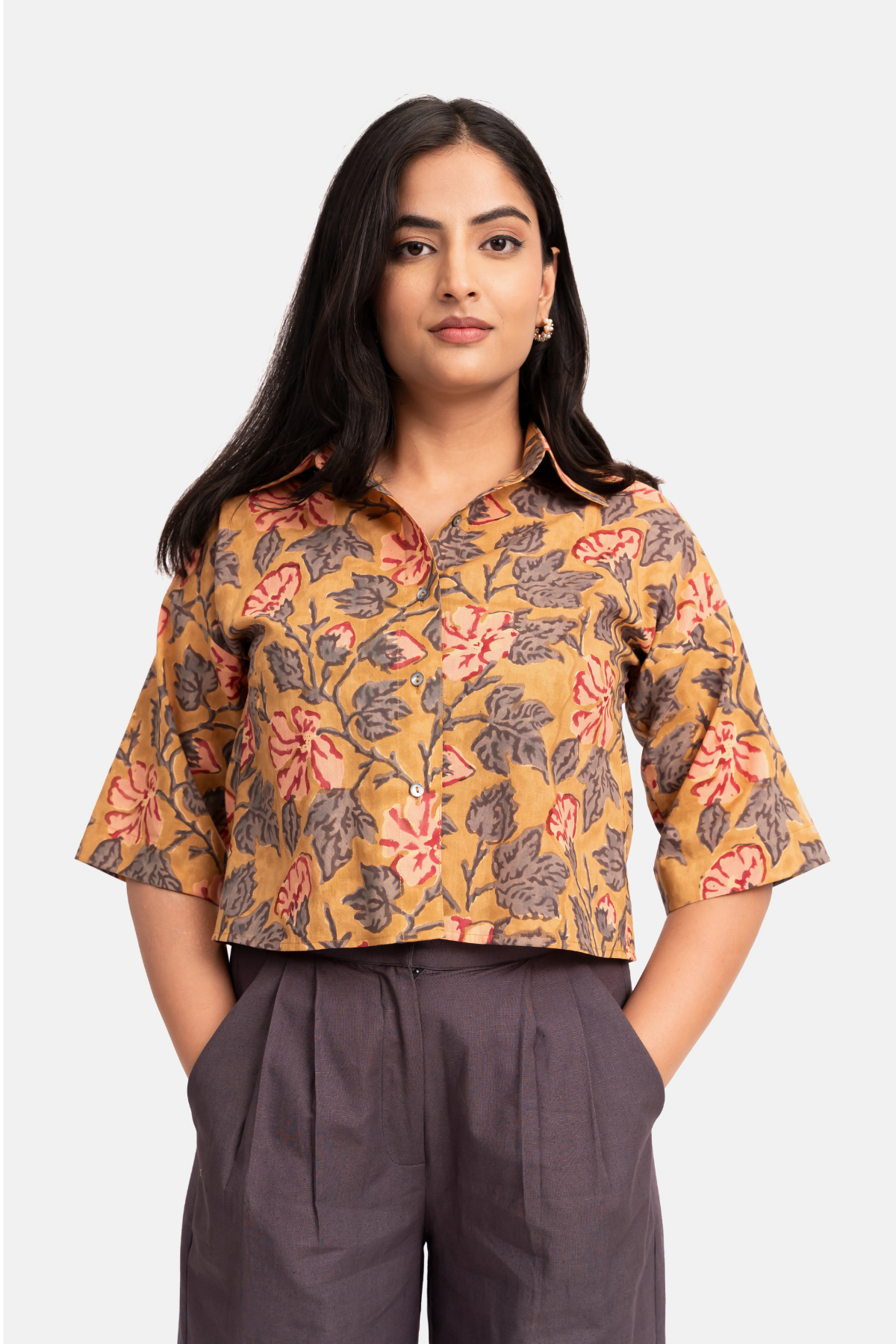A woman modeling a mustard yellow short-sleeve button-up shirt with a pink and gray floral block print, paired with dark gray trousers, against a light gray background.