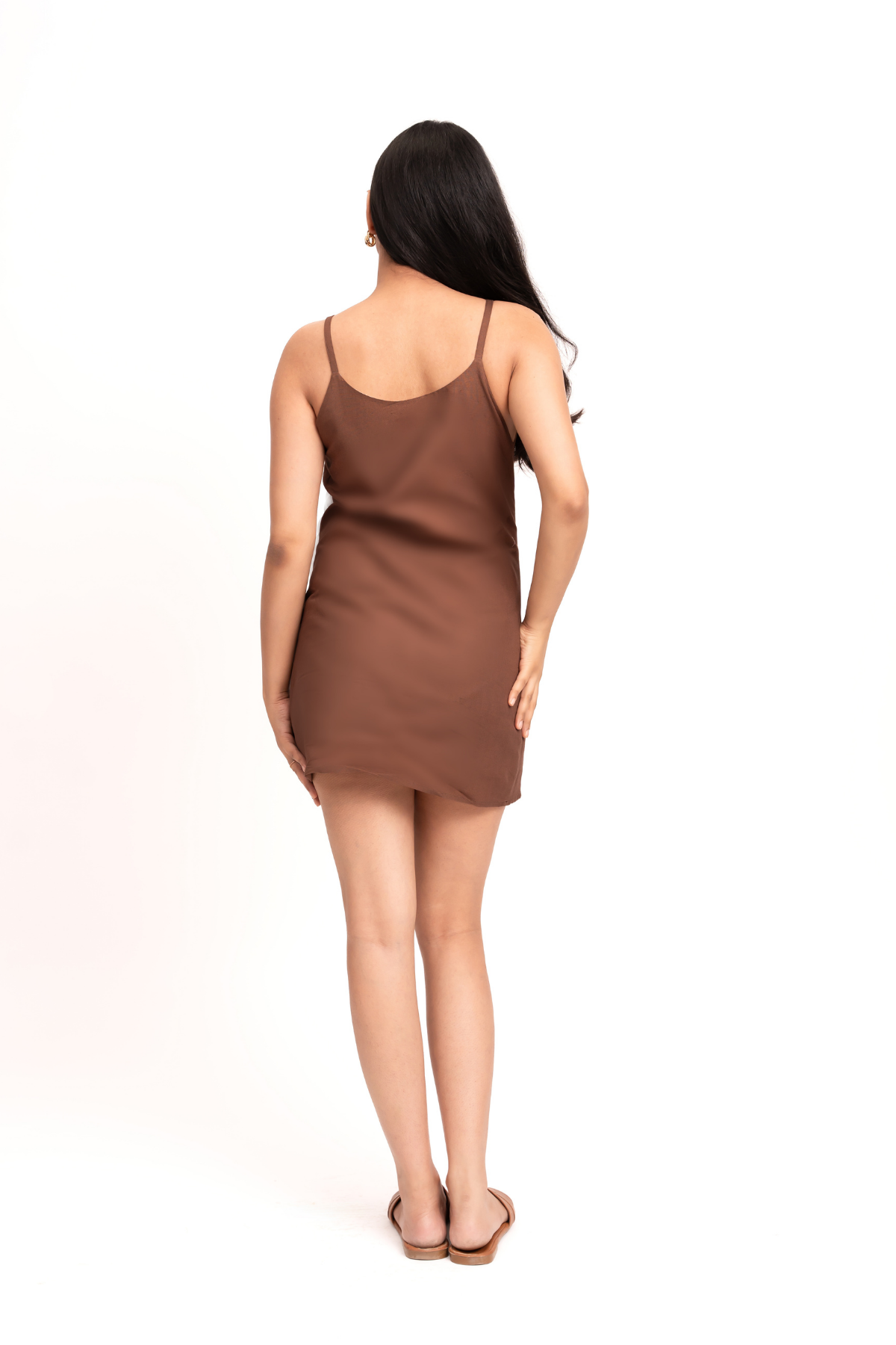 A woman viewed from behind wearing a brown spaghetti-strap mini dress and flat sandals, standing against a plain white studio background.