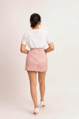 A woman is shown from behind wearing a white t-shirt and a pink and white vertically striped mini skirt, paired with beige heels, against a plain light background.