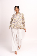 A woman modeling a beige and white printed long-sleeve blouse with a high collar and flared cuffs, paired with white wide-leg pants, against a plain white studio background.