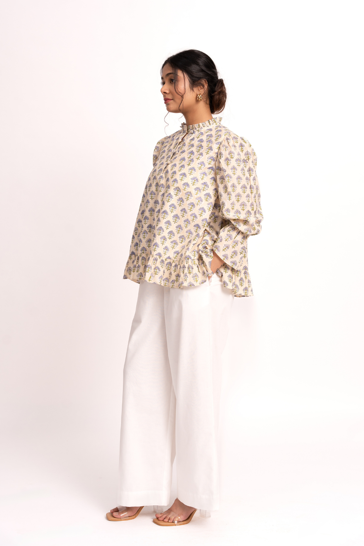 A woman modeling a beige long-sleeve blouse with a blue and yellow floral block print, paired with white wide-leg trousers and brown sandals, photographed against a seamless white studio background.