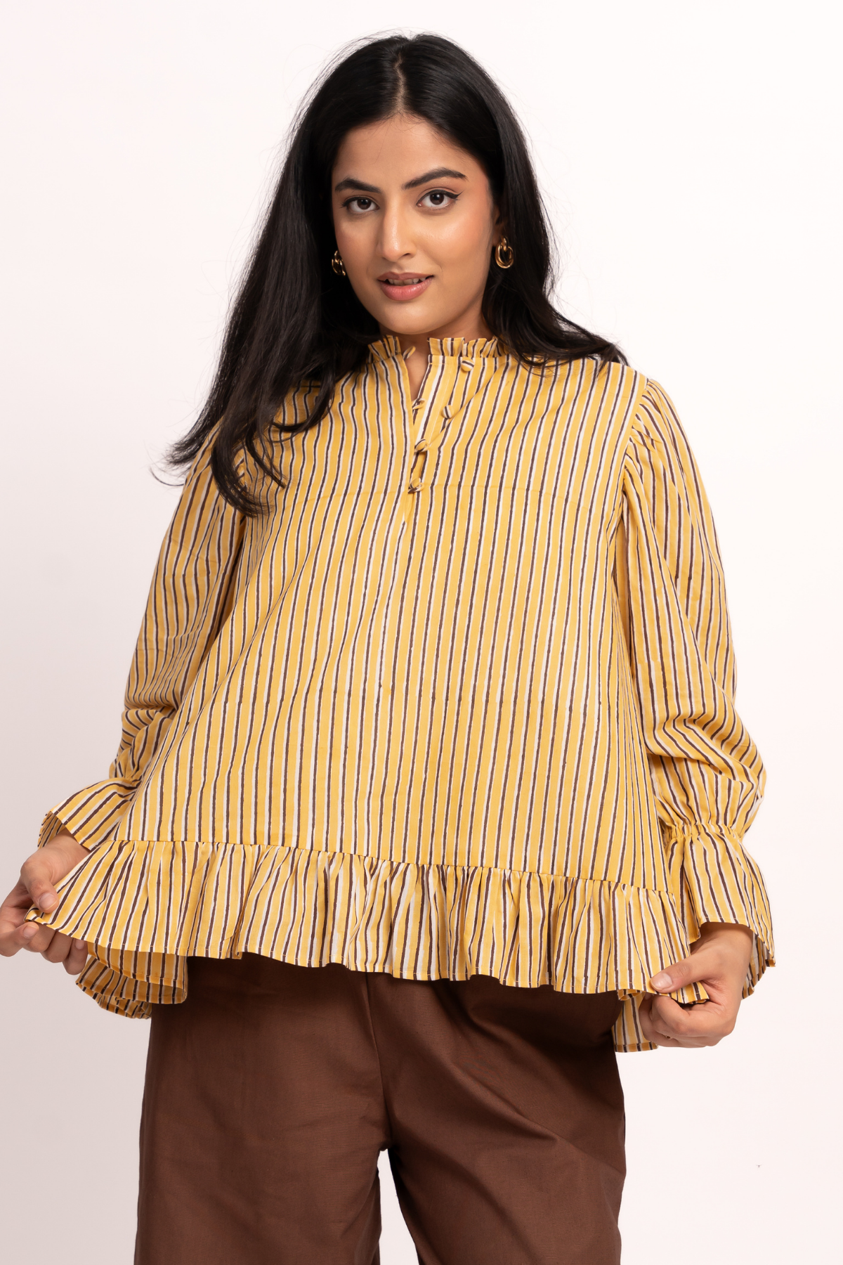 A woman modeling a mustard yellow and blue vertically striped long-sleeve blouse with a ruffled hem and gathered sleeves, paired with brown pants, against a plain white studio background.