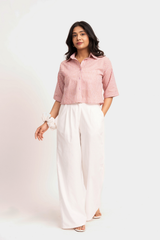 A woman modeling a pink and white vertically striped short-sleeve button-up shirt paired with white wide-leg trousers, standing against a plain light background.
