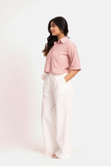 A woman modeling a pink and white vertically striped short-sleeve button-up shirt paired with white wide-leg trousers, standing against a plain off-white studio background.