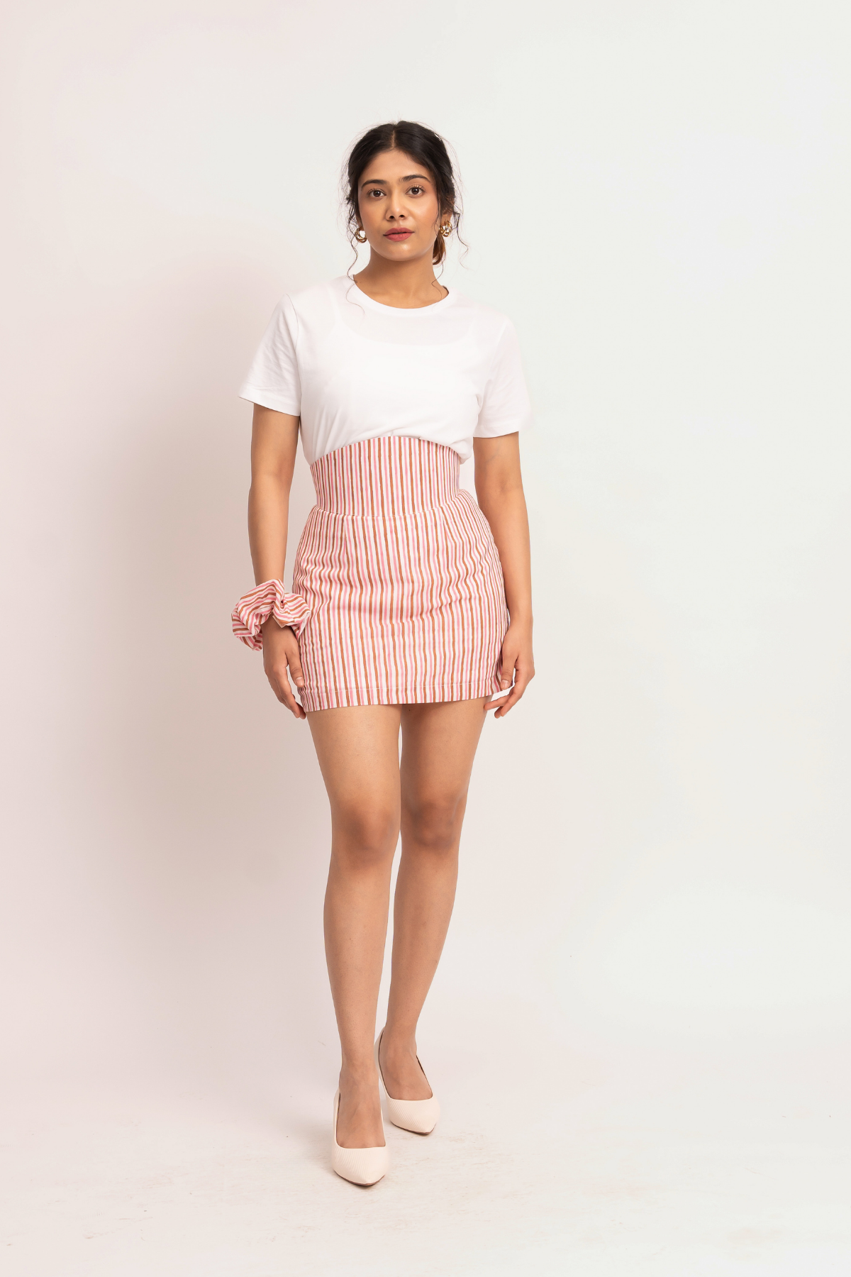 A woman modeling a white short-sleeve t-shirt and a pink and white vertically striped high-waisted mini skirt, paired with a matching pink scrunchie and beige heels, against a plain white studio background.