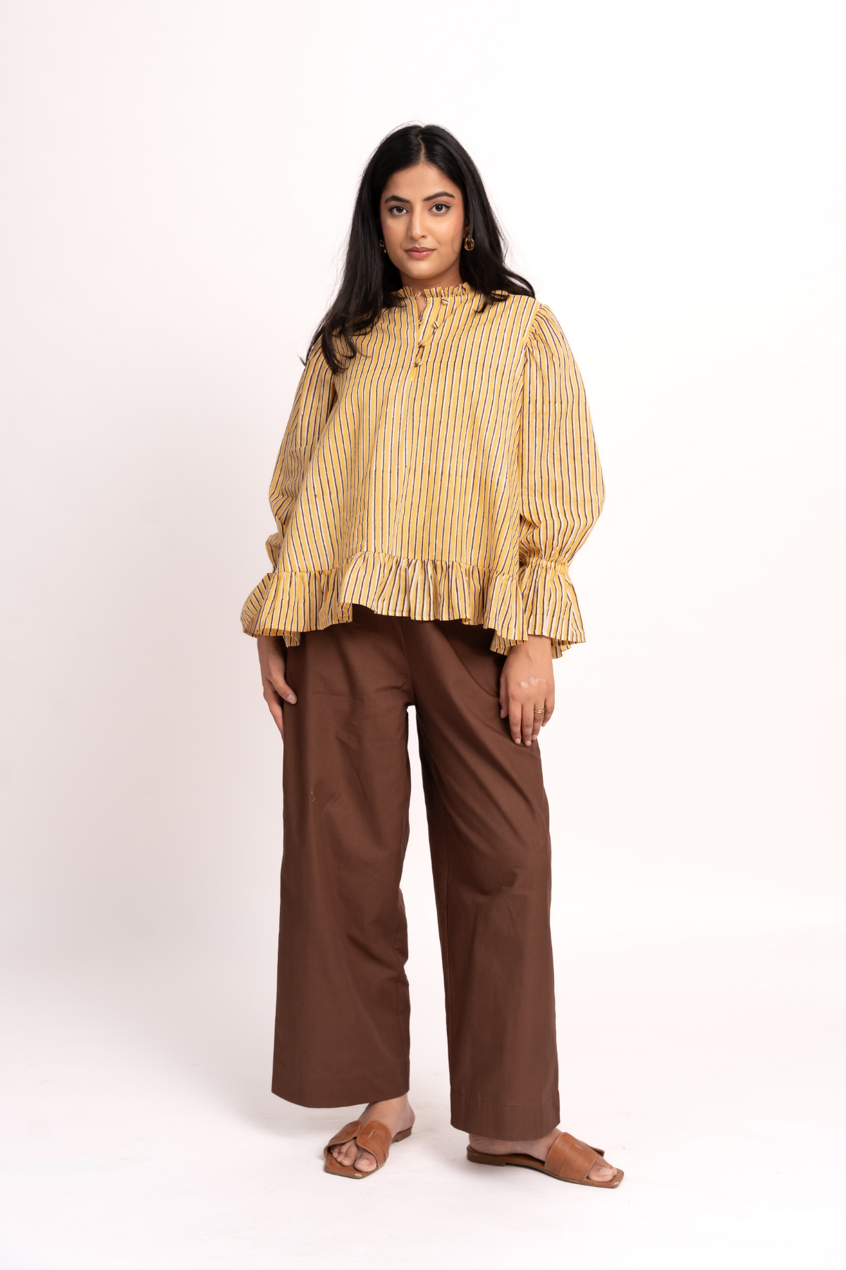 A woman modeling a yellow and brown vertically striped long-sleeve blouse with a ruffled hem, paired with brown wide-leg trousers and brown flat sandals, against a plain white studio background.