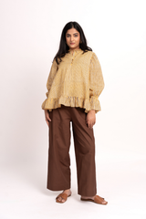 A woman modeling a yellow and brown vertically striped long-sleeve blouse with a ruffled hem, paired with brown wide-leg trousers and brown flat sandals, against a plain white studio background.