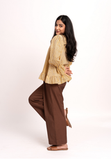 A woman posing in a studio wearing a beige and yellow striped long-sleeve blouse with ruffled cuffs and wide-leg brown trousers, photographed against a clean white background.