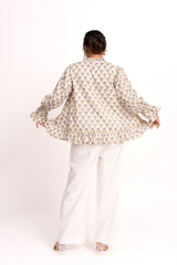 A woman shown from behind wearing a cream-colored long-sleeve blouse with a blue and yellow floral pattern and white wide-leg trousers, standing against a plain white studio background.