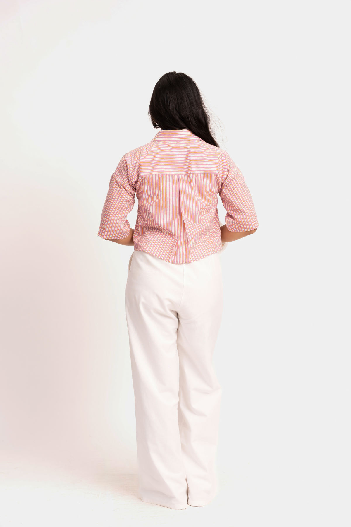 A woman viewed from behind wearing a pink and white vertically striped short-sleeve button-up shirt and off-white wide-leg trousers, standing against a plain light background.