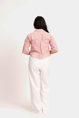 A woman viewed from behind wearing a pink and white vertically striped short-sleeve button-up shirt and off-white wide-leg trousers, standing against a plain light background.