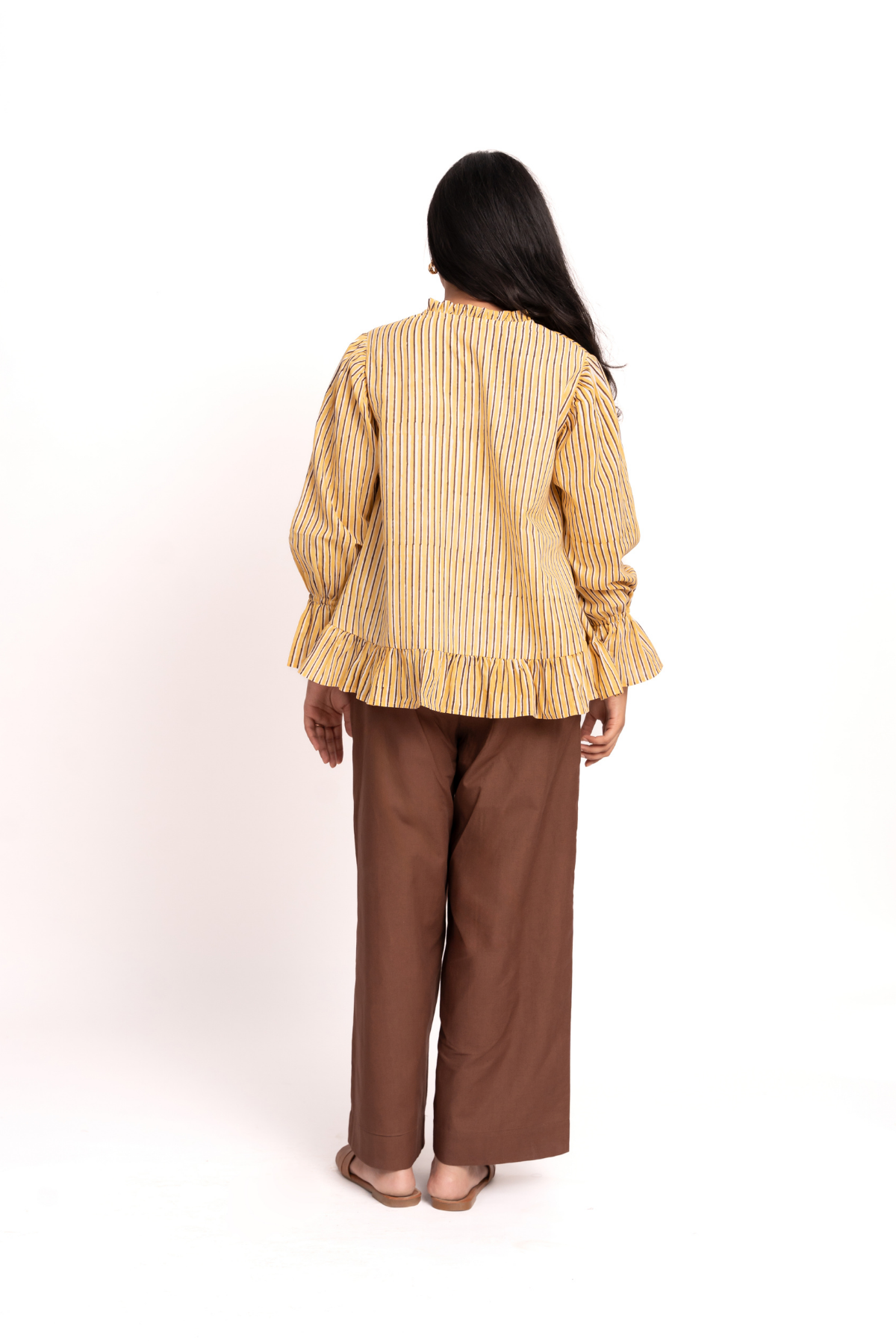 A woman viewed from behind wearing a yellow and white vertically striped long-sleeve blouse with ruffled cuffs and hem, paired with brown wide-leg pants, against a plain white studio background.