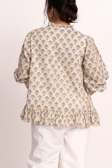 A woman viewed from the back wearing a cream-colored blouse with a repeating blue and yellow floral block print, paired with white pants, against a plain off-white background.