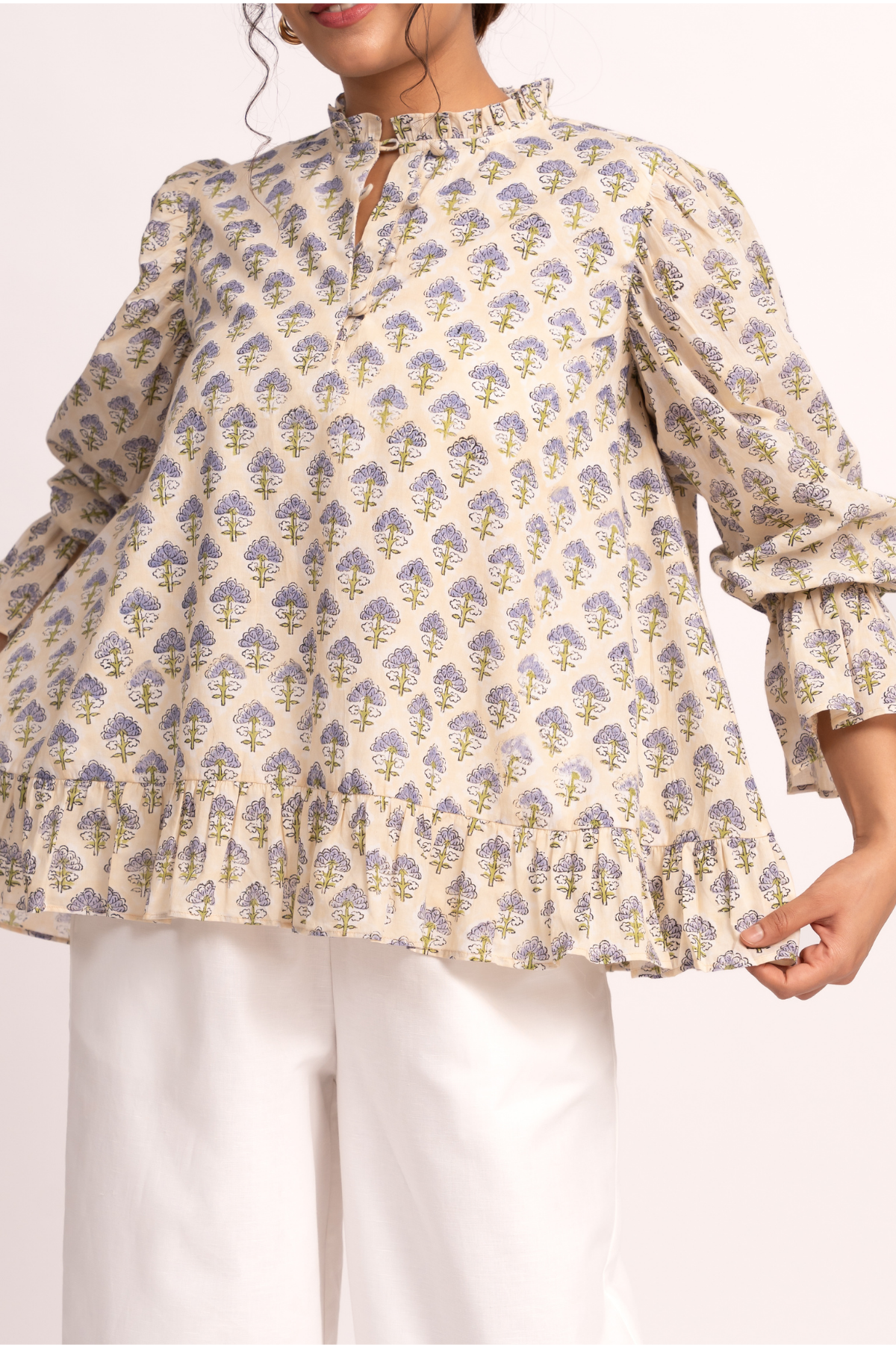 A woman wearing a cream-colored blouse with a blue and yellow floral block print, featuring a keyhole neckline, ruffled collar, and voluminous sleeves with ruffled cuffs.