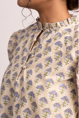 Close-up of a person wearing a cream-colored blouse with a repeating blue and green floral block print pattern and a ruffled high neckline.