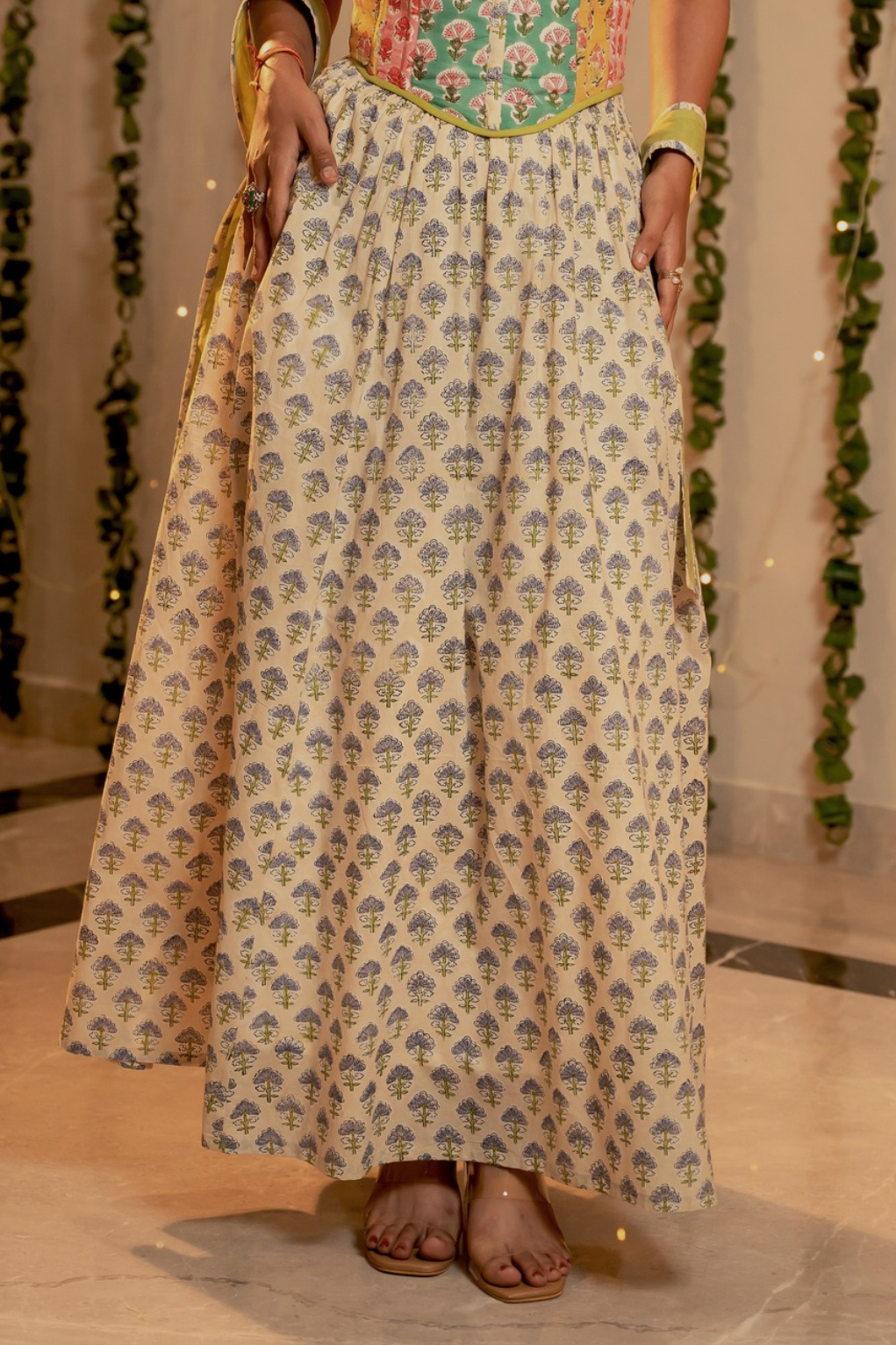 A woman wearing a long cream-colored skirt with a blue and green floral block print pattern, paired with a colorful embroidered bodice and sandals, standing in an indoor setting with decorative greenery in the background.