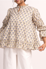 A woman wearing a cream-colored blouse with a blue and yellow floral block print, featuring a keyhole neckline, ruffled collar, and voluminous sleeves with ruffled cuffs.
