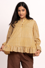 A woman modeling a mustard yellow and blue vertically striped long-sleeve blouse with a ruffled hem and gathered sleeves, paired with brown pants, against a plain white studio background.